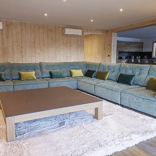 Grand salon de chalet confortable et lumineux, grand canapé d'angle, tapis, table basse, ameublement bois. Coin cuisine