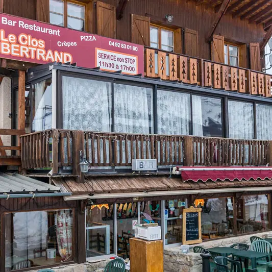 Terrasse extérieure du restaurant, tables et chaises, et bâtiment principal, en pierre, sur deux étages. Panneau au 2e étage : Bar Restaurant Le Clos Bertrand