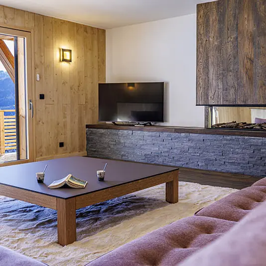 Grand et lumineux séjour, table basse, canapé, TV, ameublement bois et pierre, porte-fenêtre donnant sur un balcon en bois et paysage enneigé