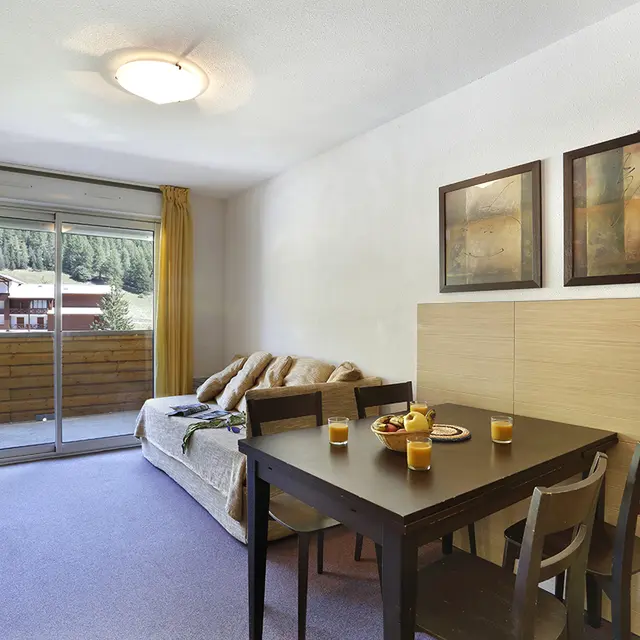 Séjour lumineux d'appartement avec canapé, meuble TV, TV, tabl à manger, porte-fenêtre donnant sur un balcon, vue sur la station