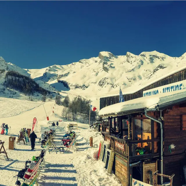 Vue de côté du restaurant d'altitude, bâtiment en bois, terrasse extérieure avec chaises longues le long des pistes enneigées, panneau Altiplano - Altitude. Montagnes en arrière-plan