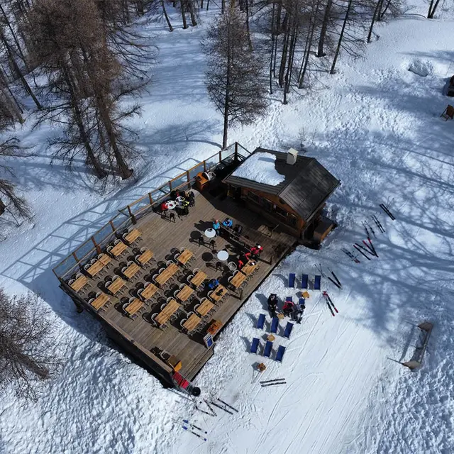 Vue du restaurant par drone, chalet et terrasse en bois au milieu des pistes et de la forêt, tables et chaises, chaises longues