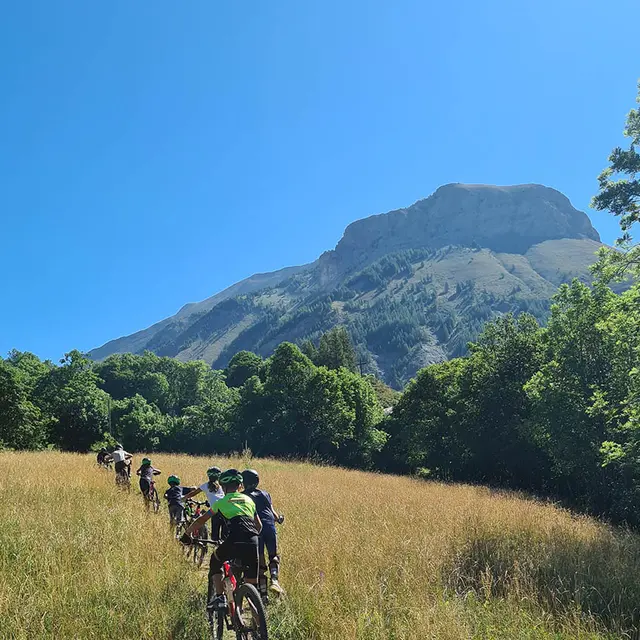 VTTistes progressant sur le plateau de Montgros, prairie en été entourée d'arbres, montagne de Montgros en arrière-plan