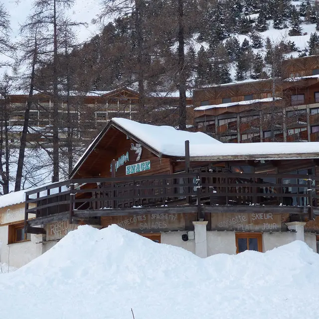 Vue d'ensemble hivernale du restaurant en bois, situé devant des immeubles de la station de ski, en haut d'un bâtiment sur deux niveaux, terrasse. Panneau Sympa Snack sur la devanture. Montagnes enneigées en arrière-plan.
