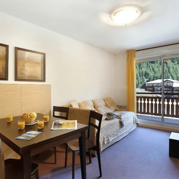 Séjour lumineux d'appartement avec canapé, meuble TV, TV, tabl à manger, porte-fenêtre donnant sur un balcon, vue sur la station