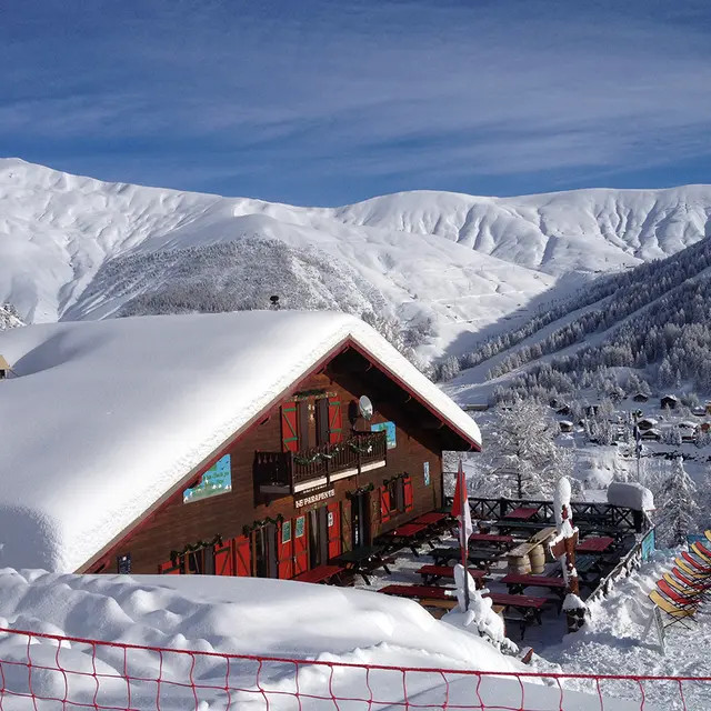 Vue du restaurant, chalet en bois, avec sa terrasse, tables et chaises, chaises lonuges. Montagnes enneigées tout autour et en arrière-plan