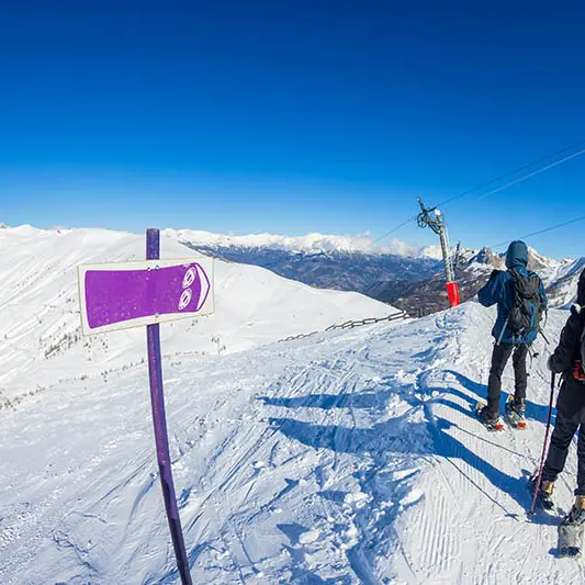 3 randonneurs en raquette au sommet d'un sentier de randonnée se situant près d'un téléski visible, grande surfaces enneigées et montagnes