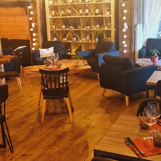 Intérieur du restaurant, ambiance chaleureuse avec fauteuils confortables, intérieur bois, tables en bois, bouteilles de vin en présentoir, porte-fenêtre donnant sur la terrasse de l'établissement